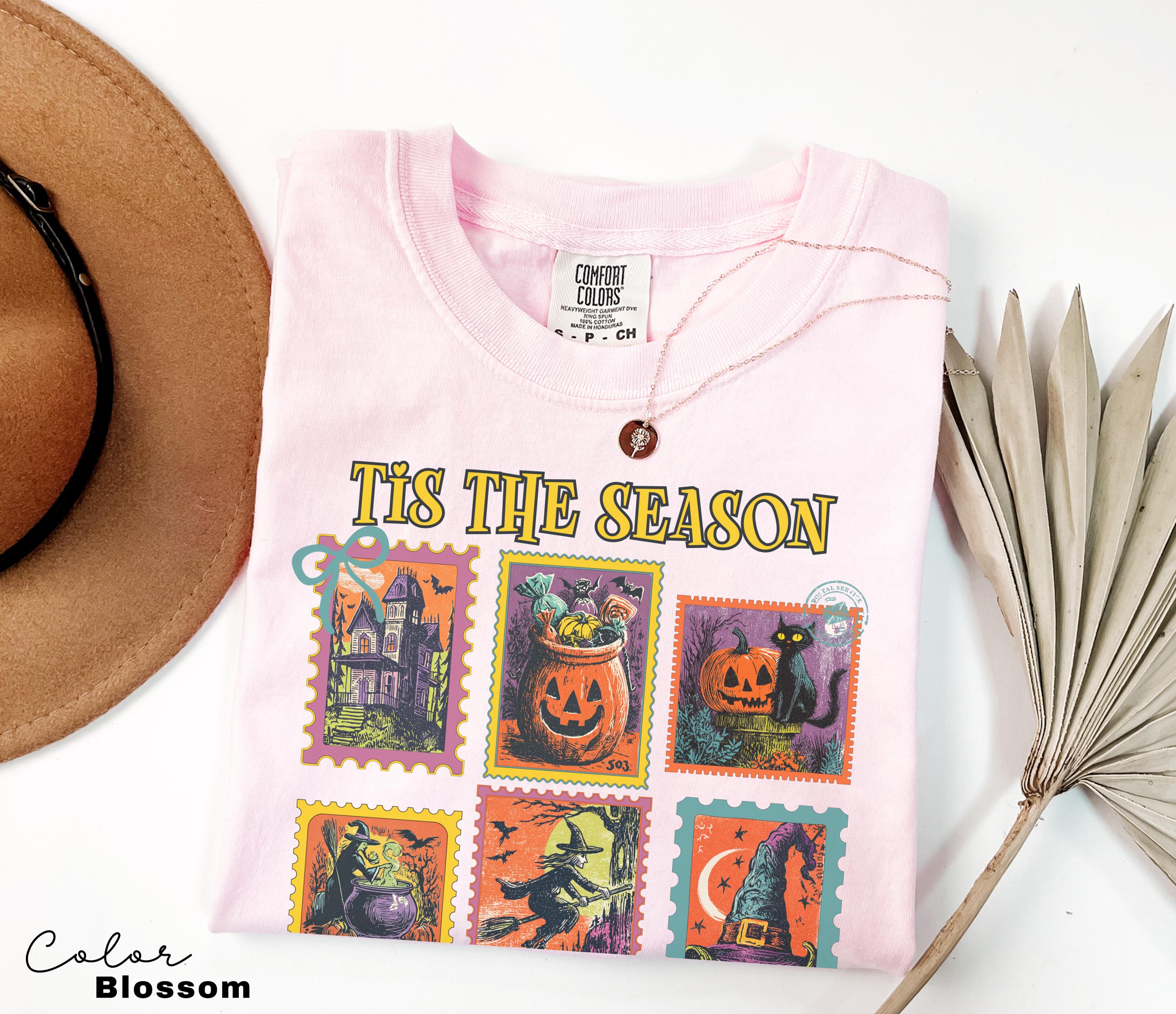 A pink t-shirt with a Halloween-themed design, including illustrations of pumpkins, bats, and a castle, along with the text "Tis the Season". The t-shirt is laid out on a white surface, accompanied by a brown hat, a plant, and a fan.