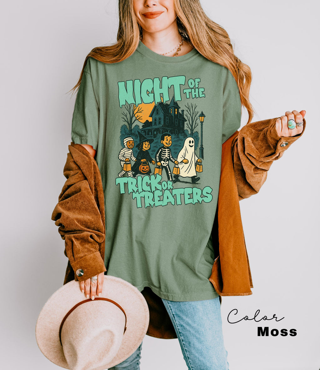 A woman wearing a green t-shirt with a Halloween-themed graphic and holding a hat stands against a white background.