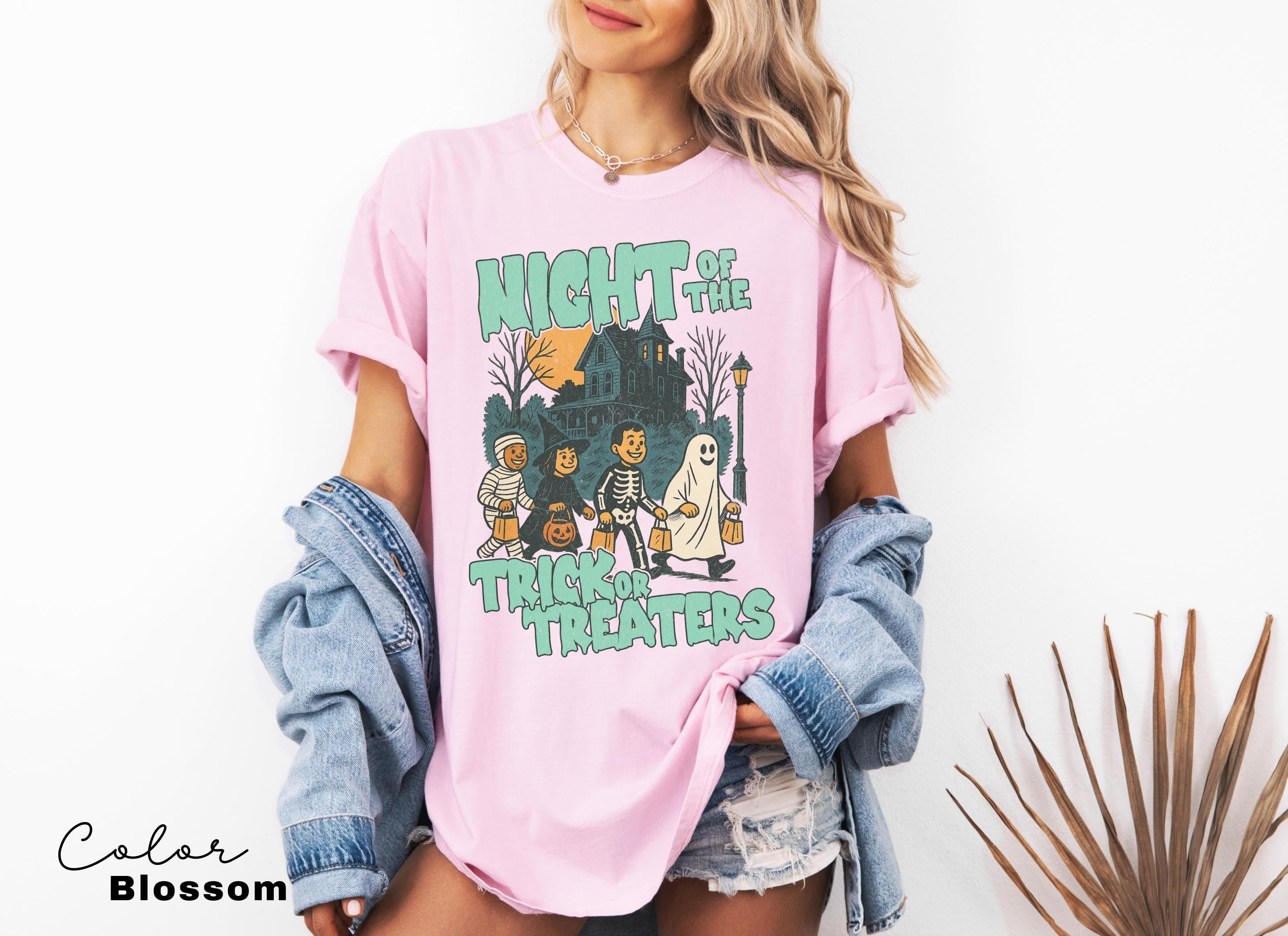 A woman wearing a pink t-shirt with a graphic design of a skeleton and ghost, along with the text "Night of the Trick Treats", standing in front of a plant.