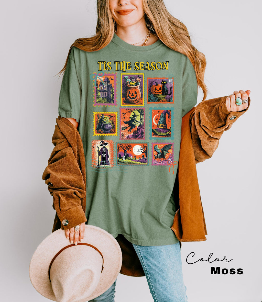 A woman wearing a green t-shirt with a graphic of Halloween-themed illustrations and holding a beige hat.