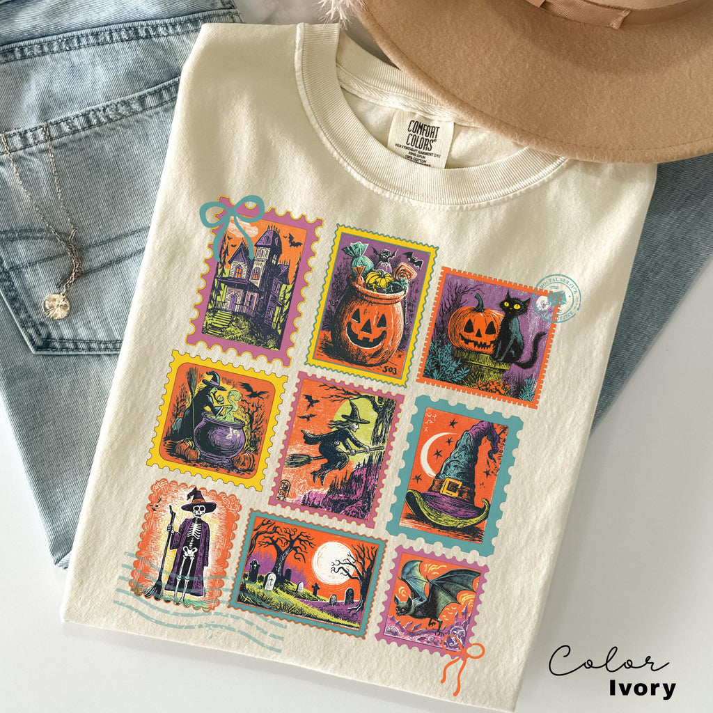 A white t-shirt with a colorful design of Halloween-themed stamps, including a castle, pumpkin, witch, cat, and other spooky and festive illustrations.