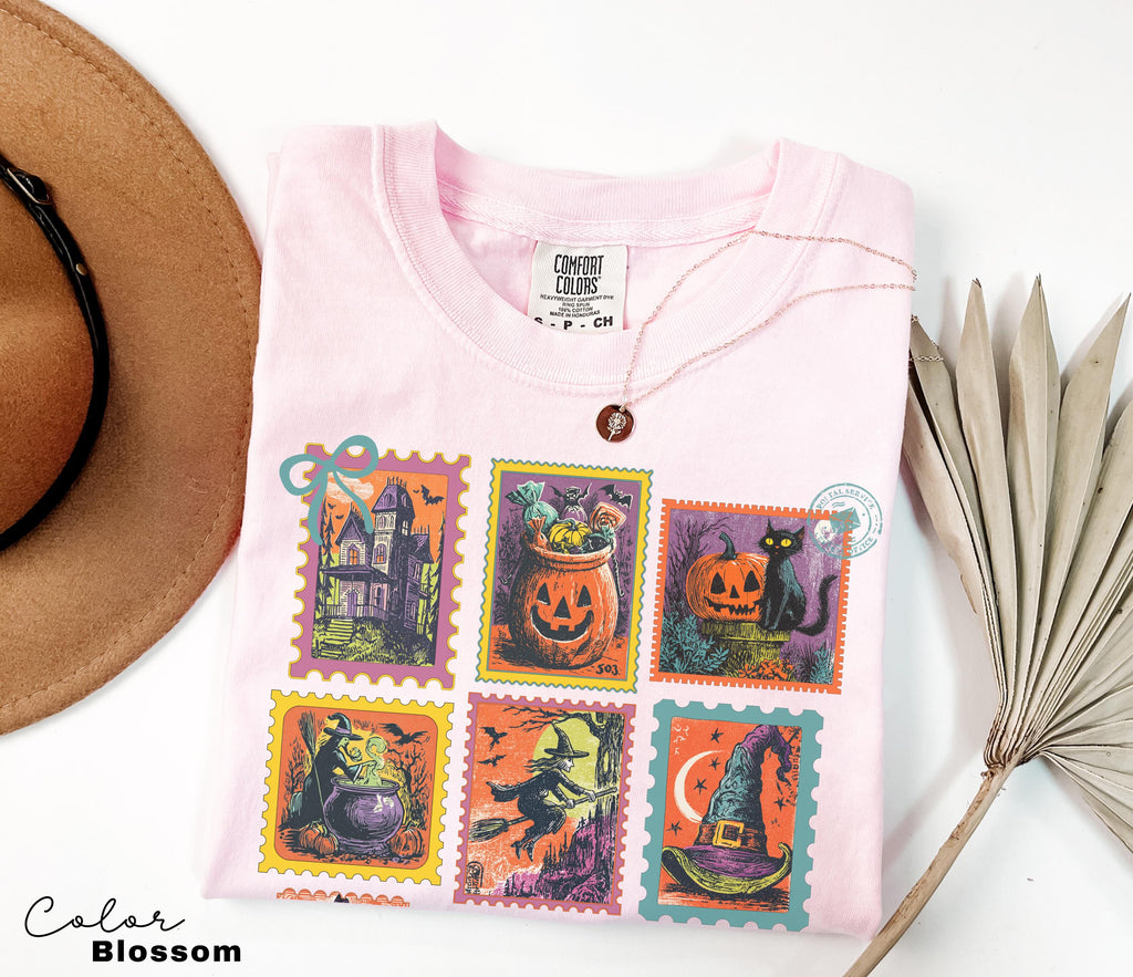 A pink t-shirt with a Halloween-themed design, including a witch, jack-o-lantern, and other spooky illustrations, is displayed alongside a brown hat and a palm frond.