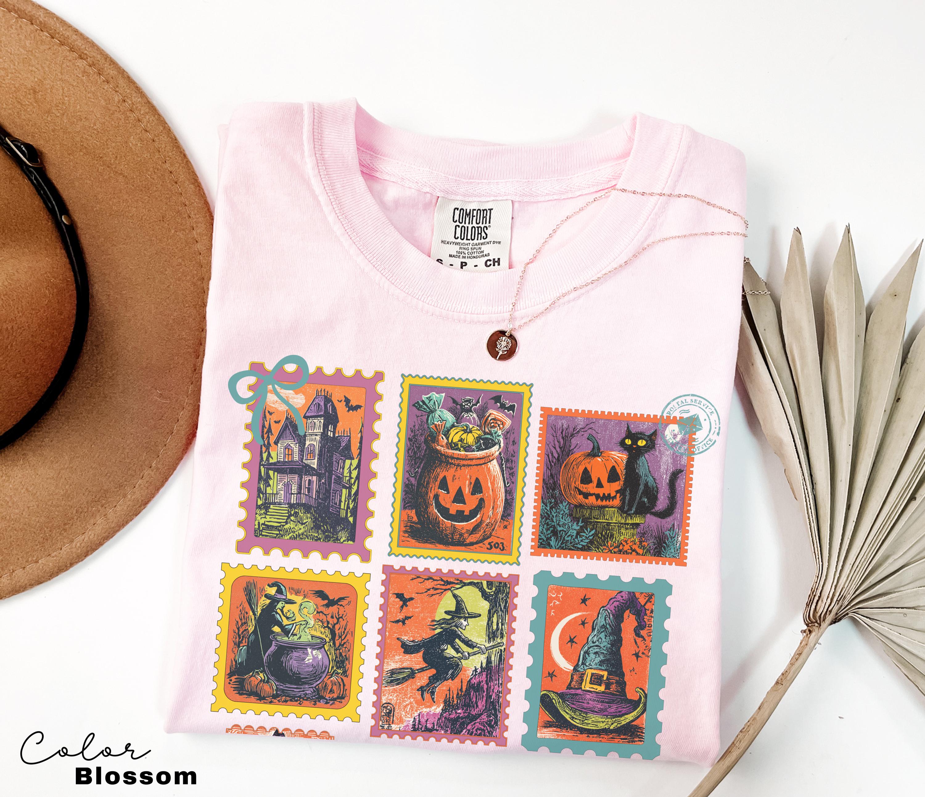 A pink t-shirt with a Halloween-themed design, including a witch, jack-o-lantern, and other spooky illustrations, is displayed alongside a brown hat and a palm frond.