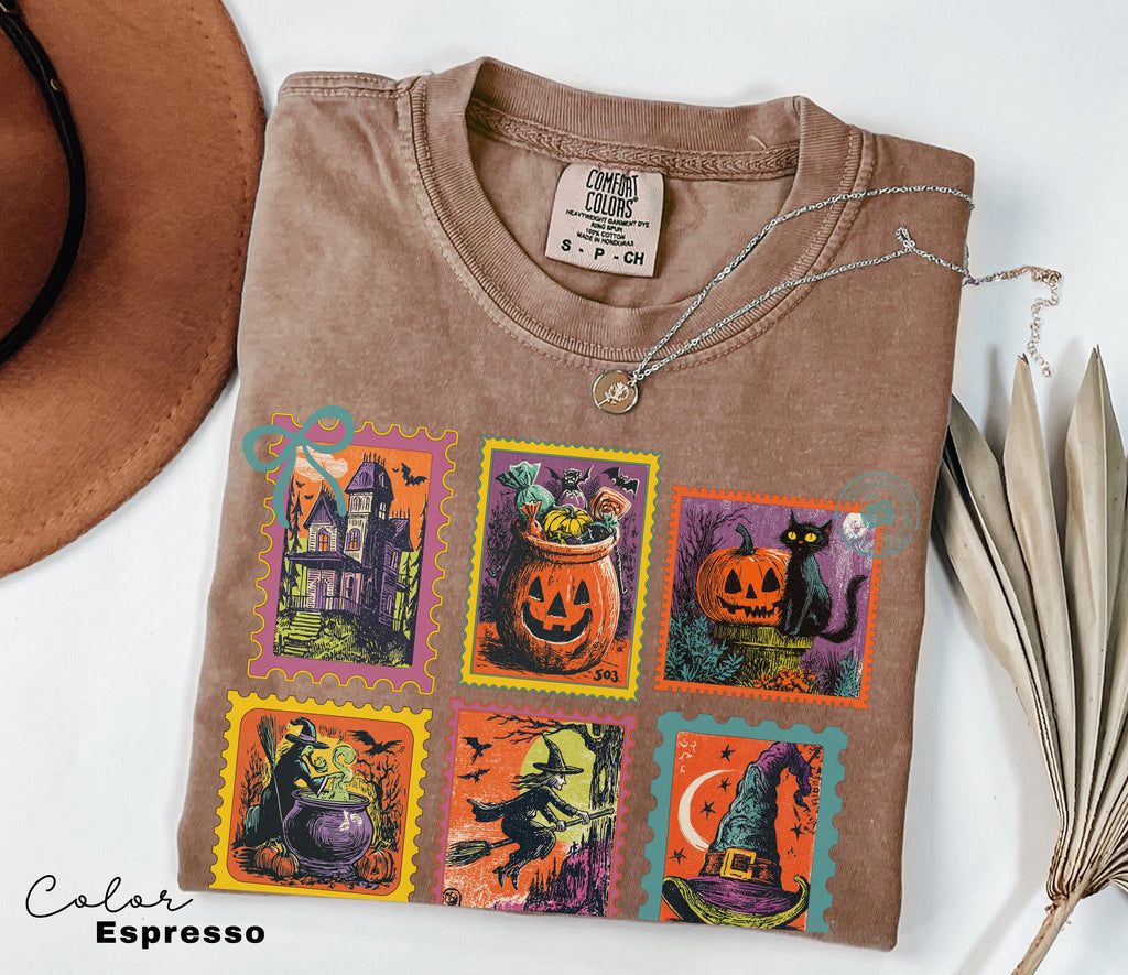 A t-shirt with a Halloween-themed design, including a pumpkin, a witch, a castle, a jack-o-lantern, a broomstick, and a witch's hat, is displayed alongside a hat and some leaves.