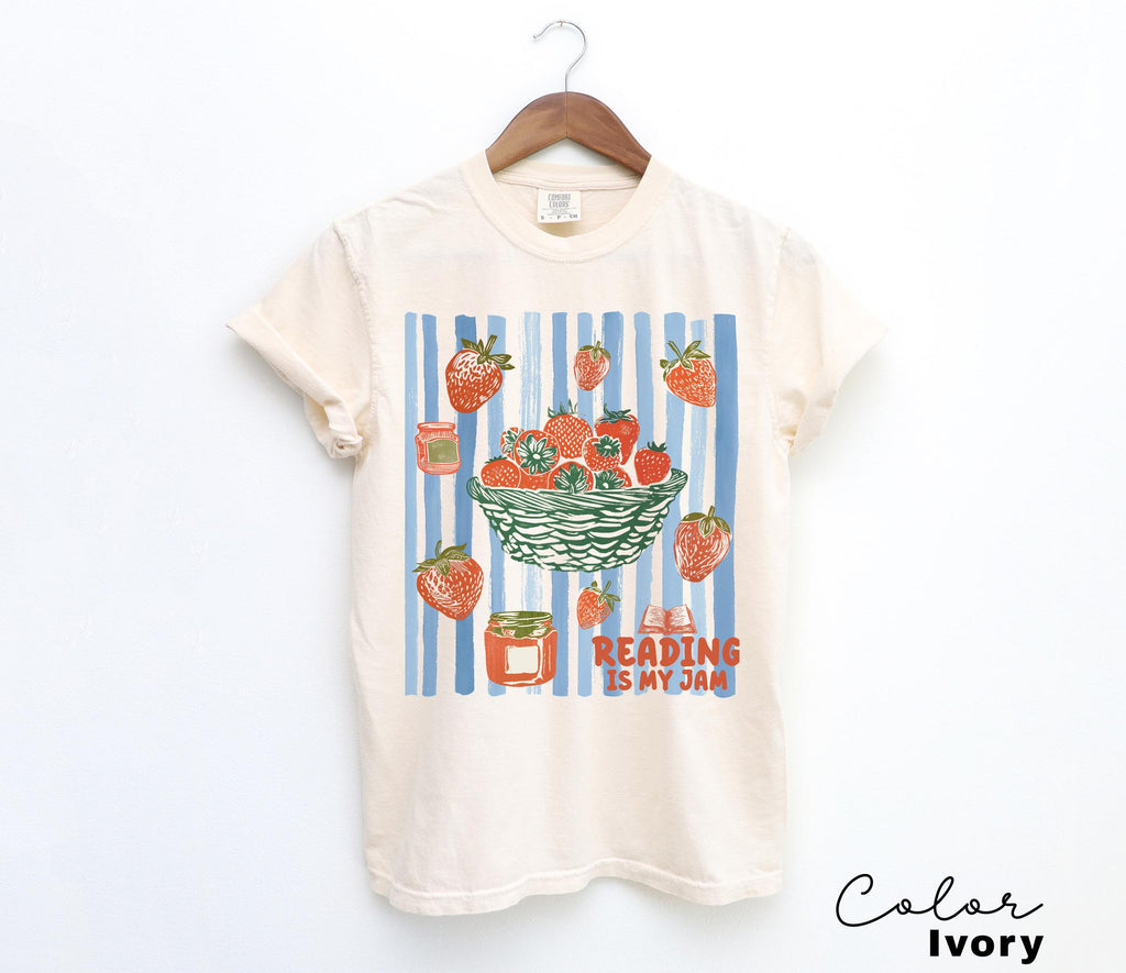 May include: Ivory-colored t-shirt with a strawberry-themed graphic. The design features a basket of strawberries, scattered berries, and jars of jam against a blue and white striped background. The text "Reading is my jam" is also included.