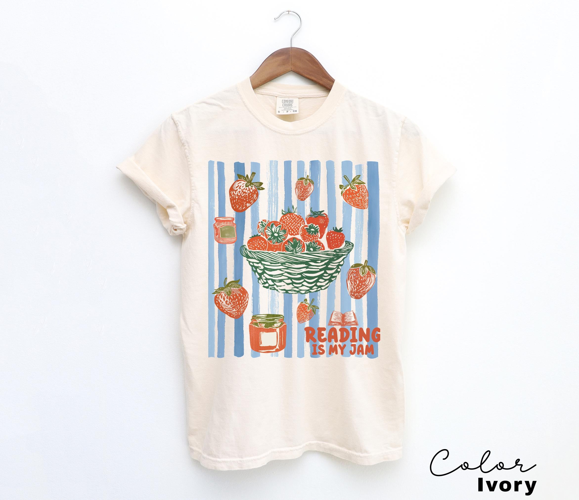 May include: Ivory-colored t-shirt with a strawberry-themed graphic. The design features a basket of strawberries, scattered berries, and jars of jam against a blue and white striped background. The text "Reading is my jam" is also included.